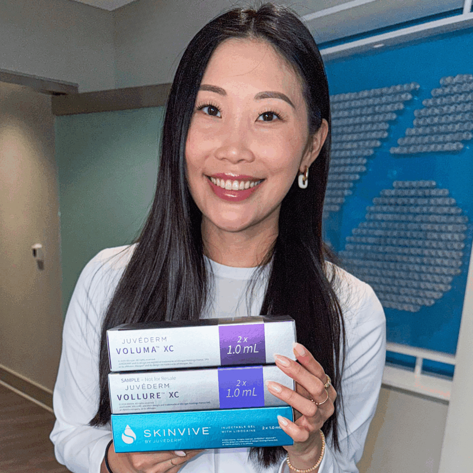 A woman smiling in a clinic holding Juvéderm Voluma, Vollure and Skinvive dermal filler boxes.