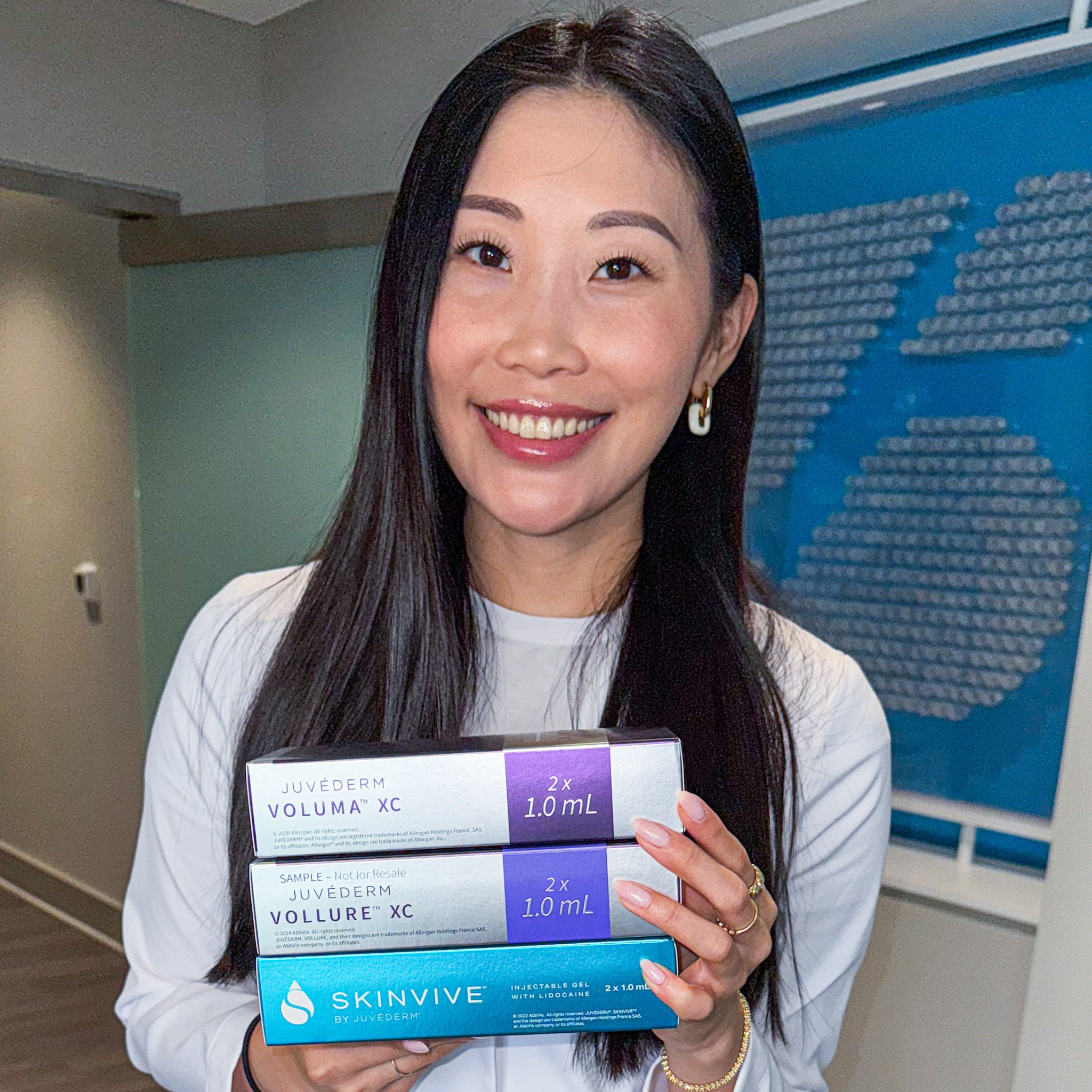 A woman smiling in a clinic holding Juvéderm Voluma, Vollure and Skinvive dermal filler boxes.