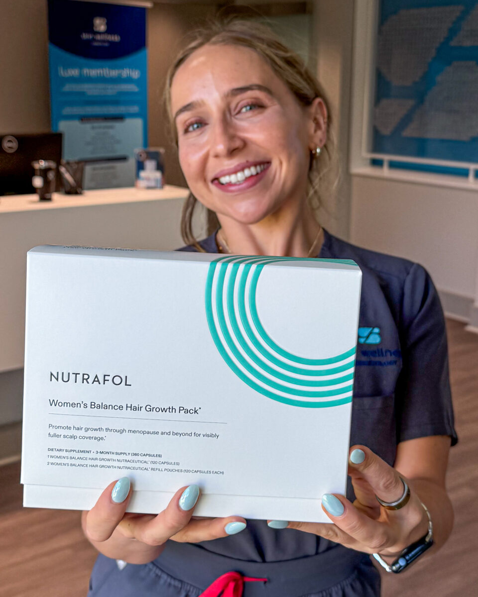 A woman at a dermatology clinic holding a Nutrafol Women's Balance Hair Growth Pack, smiling