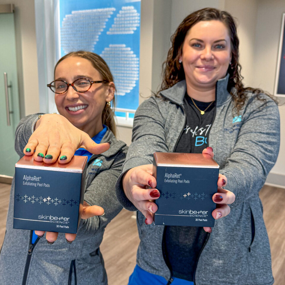 Two clinic staff smiling, holding SkinBetter Science AlphaRet exfoliating peel pads boxes toward camera