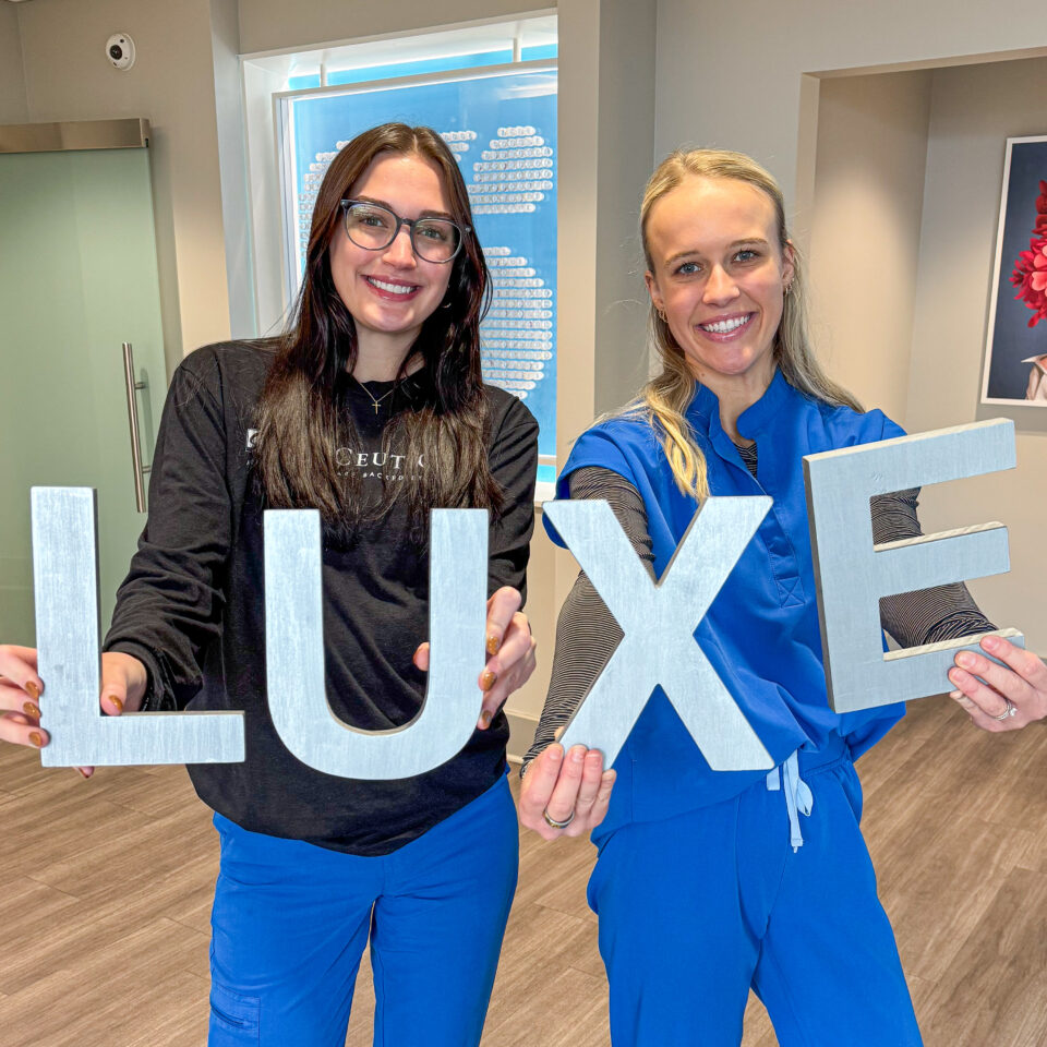 Two smiling staff in blue scrubs holding large LUXE letters at a Birmingham dermatology clinic reception