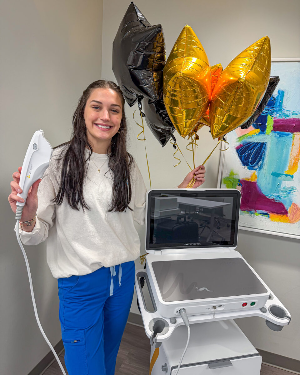 A woman holding an Ultherapy handpiece and gold balloons beside an aesthetic treatment machine in a Birmingham clinic.