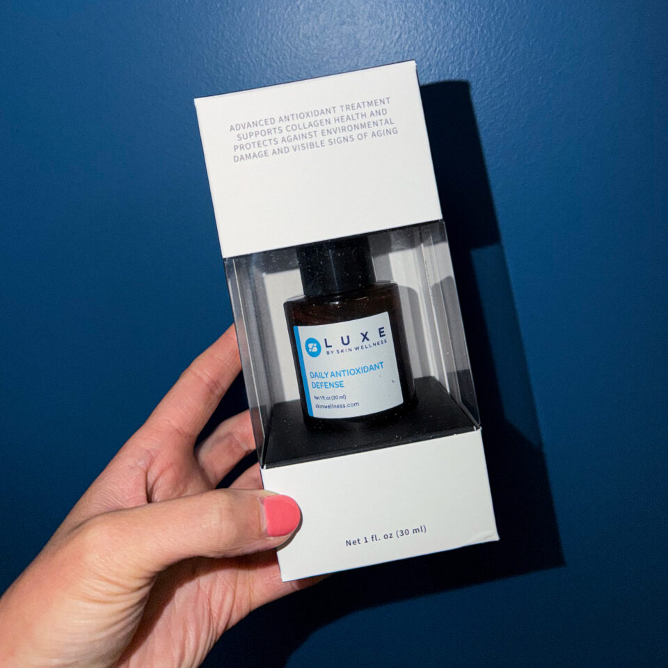 A person holding a boxed antioxidant serum (Luxe by Skin Wellness) against a blue background