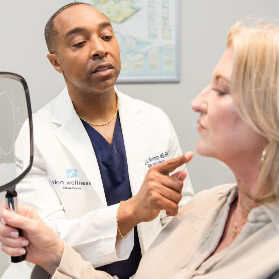 Dermatologist examines a patient's chin during cosmetic consultation as patient holds a mirror.