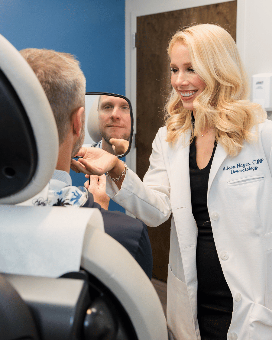 A woman in a white coat shows a male patient his reflection in a dermatology clinic, smiling and discussing treatment