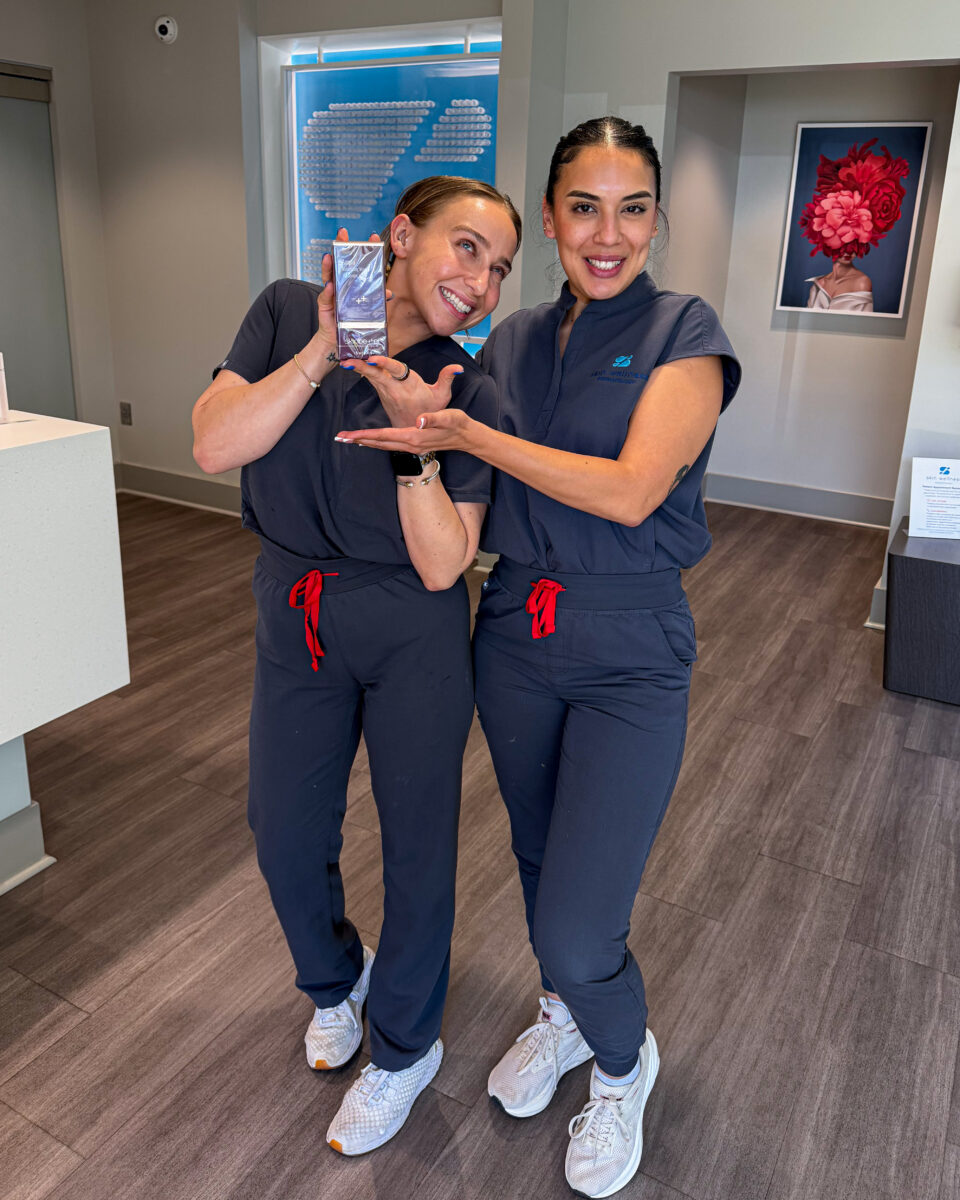 Two clinic staff in scrubs smiling and presenting a skincare product at Birmingham dermatology clinic.