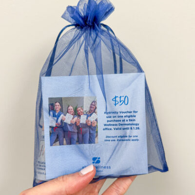 A hand holding a blue organza gift bag with a $50 Skin Wellness Dermatology voucher and promo materials