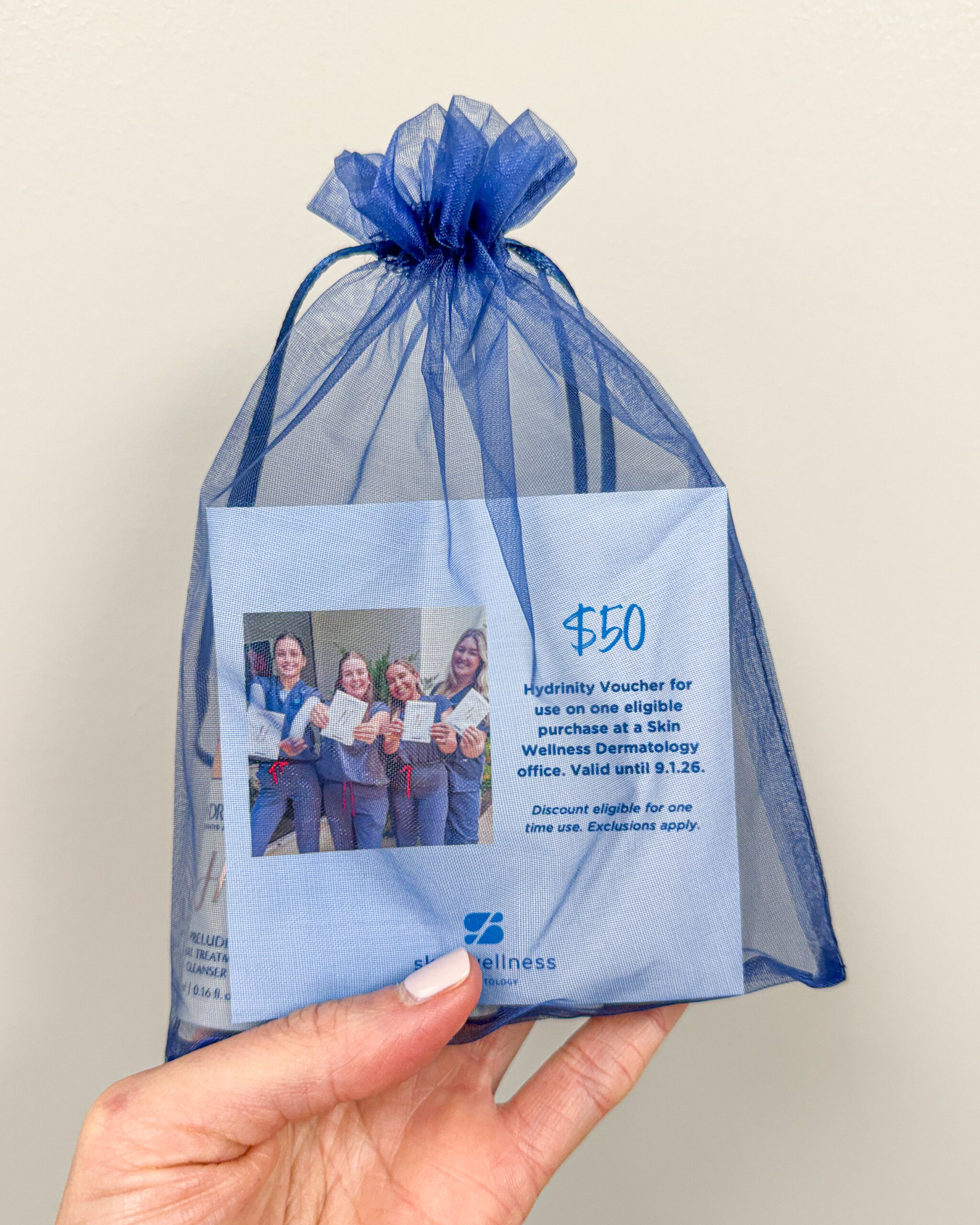 A hand holding a blue organza gift bag with a $50 Skin Wellness Dermatology voucher and promo materials