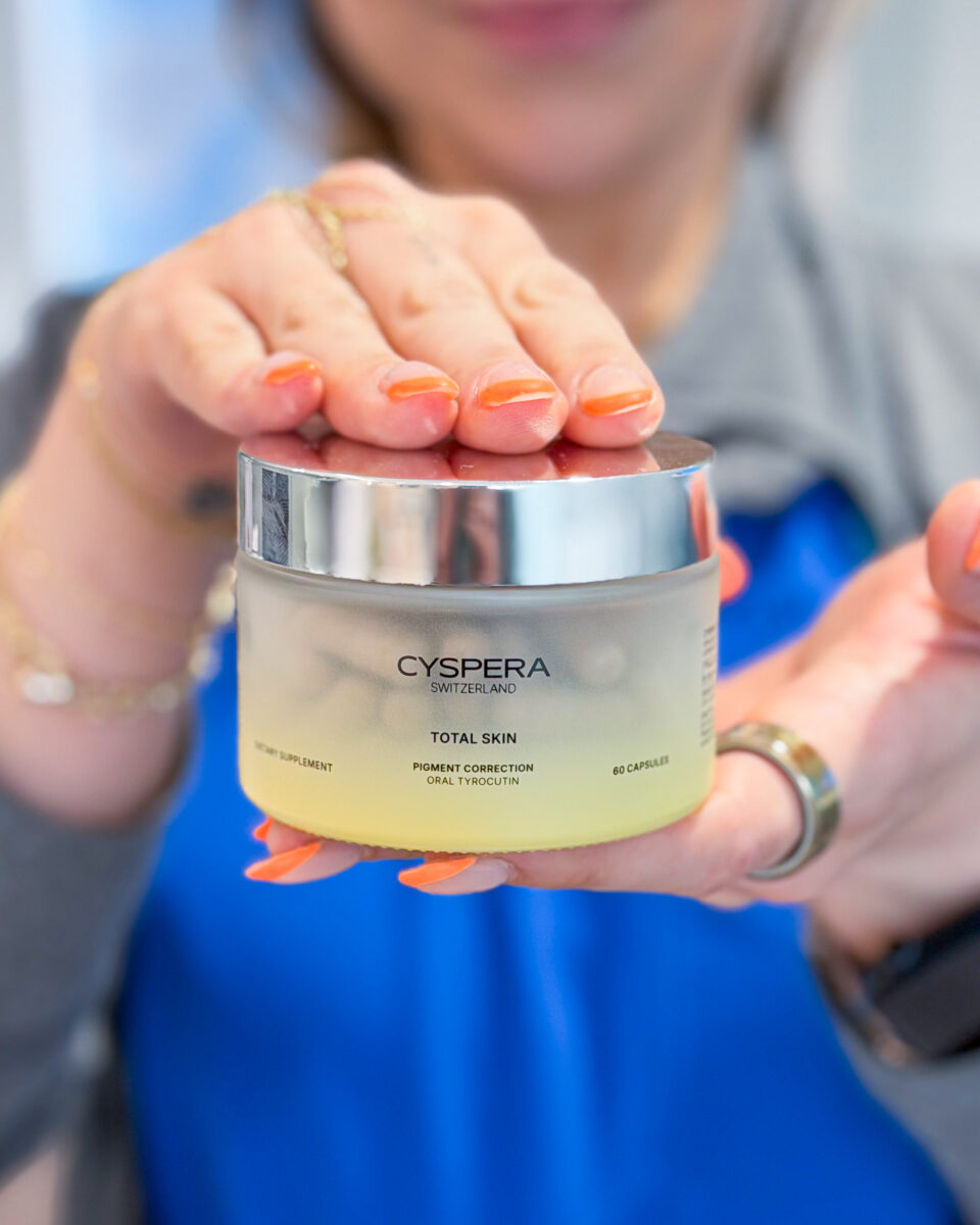 A person with orange manicure holding a jar of Cyspera Total Skin pigment correction skincare