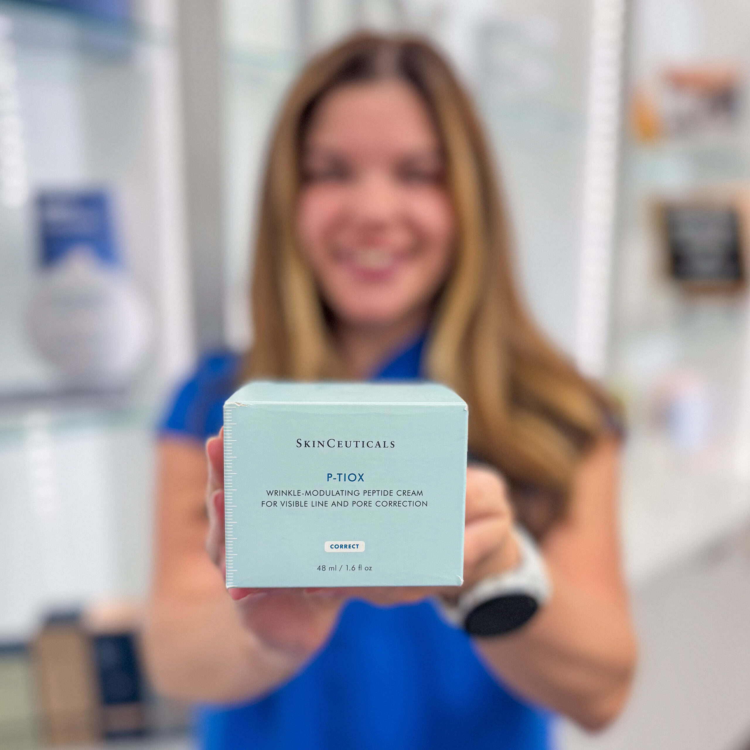 A woman in a dermatology clinic holding a SkinCeuticals P-Tox box for wrinkle-modulating peptide cream.